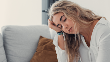 світловолоса жінка тримається за голову на дивані як ілюстрація до матеріалу про допомогу при сильному головному болю у клініці Медонія Прайм