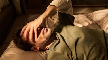 чоловік лежить у ліжку та тримається за голову через сильний нічний головний біль як ілюстрація до матеріалу про причини сильного головного болю в клініці Медонія Прайм