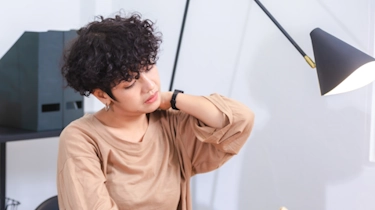 молода жінка з кучерявим волоссям тримається за шию через біль як ілюстрація до матеріалу про обстеження шийного відділу у клініці Медонія Прайм