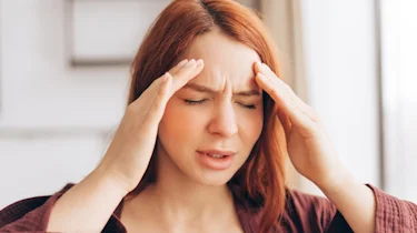 рудоволоса жінка тримається за скроні через тривалий головний біль як ілюстрація до матеріалу про консультацію при тривалому головному болю в клініці Медонія Прайм