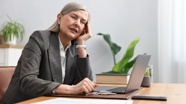 старша жінка сидить за ноутбуком і тримається за голову через головокружіння як ілюстрація до матеріалу про консультацію невролога при головокружінні у жінок в клініці Медонія Прайм