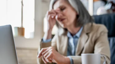 жінка за ноутбуком тримається за голову через погане самопочуття як ілюстрація до матеріалу про консультацію при головному болю та слабкості в клініці Медонія Прайм