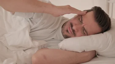 чоловік лежить у ліжку та тримає руку біля голови через сильний біль як ілюстрація до матеріалу про нічний напад кластерного головного болю в клініці Медонія Прайм