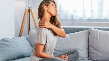 жінка з болем у шиї сидить удома як ілюстрація до матеріалу про допомогу при шийному остеохондрозі у клініці Медонія Прайм