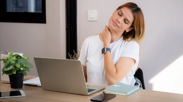 молода жінка сидить за столом і тримається за шию як ілюстрація до матеріалу про профілактику шийного остеохондрозу у клініці Медонія Прайм