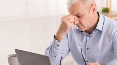 чоловік за ноутбуком тримається за чоло через сильний головний біль як ілюстрація до матеріалу про діагностику кластерного головного болю та впливу стресу в клініці Медонія Прайм